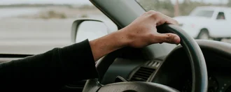 Innenansicht eines Autos auf einer Autobahn. Der linke Arm einer Person hält die obere Seite des Lenkrads fest. Im Hintergrund kann durch die Seiten- und Frontscheibe auf die Straße geblickt werden.
