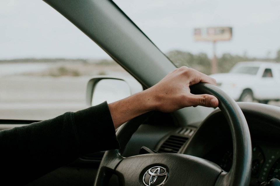 Innenansicht eines Autos auf einer Autobahn. Der linke Arm einer Person hält die obere Seite des Lenkrads fest. Im Hintergrund kann durch die Seiten- und Frontscheibe auf die Straße geblickt werden.
