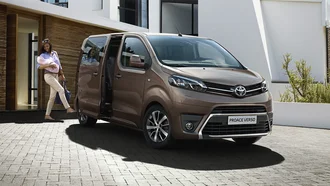 Brauner Toyota Proace Verso vor einem Haus fotografiert. Mutter mit Baby auf dem Arm läuft zum Auto