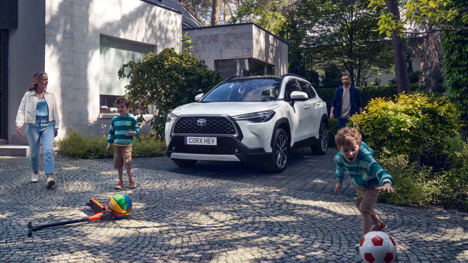 Ein Toyota Corolla Cross steht in der Einfahr eines Grundstücks umgeben von einer Familie mit Frau, Mann und zwei Kindern.